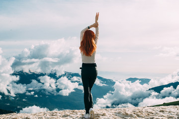 A slender long-haired ginger girl stands on the top of a snowy mountain against the background of clouds. Back view, open space for your own text - 341040545