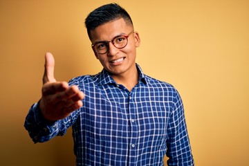 Young handsome latin man wearing casual shirt and glasses over yellow background smiling friendly offering handshake as greeting and welcoming. Successful business.