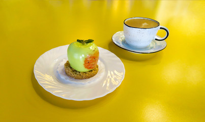 Cappuccino coffee stands in a Cup on the yellow table. Dessert cake in the form of an Apple lying on a silver platter. White napkins in a napkin holder are on the table. Dark blue background.