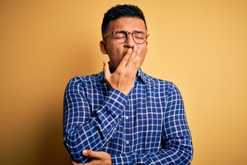 Fototapeta premium Young handsome latin man wearing casual shirt and glasses over yellow background bored yawning tired covering mouth with hand. Restless and sleepiness.