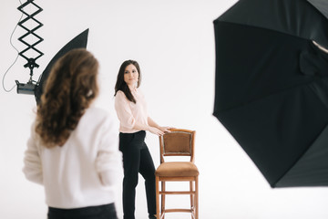 Attractive young female model posing with chair in photo studio for female photographer while she is shooting with digital camera. Concept of creative work in photo studio, backstage job.