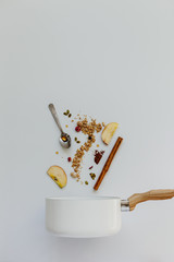 Apples, oat granola, cinnamon stick, pumpkin seeds, dried berries and syrup going into a white saucepan on a pastel white background
