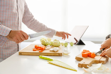 A girl's hands cut vegetables to cook while her father looks at the recipe on the tablet