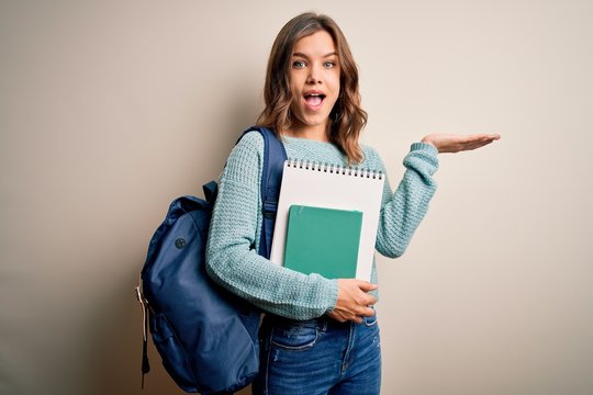 Young blonde student girl wearing backpack and books from school over isolated background very happy and excited, winner expression celebrating victory screaming with big smile and raised hands