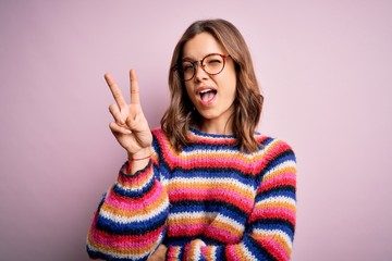 Young beautiful blonde girl wearing glasses and casual sweater over pink isolated background smiling with happy face winking at the camera doing victory sign with fingers. Number two.
