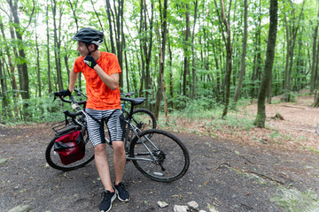 Naklejka premium Man with bicycle in full cycling equipment with helmet, orange t-shirt in green summer forest