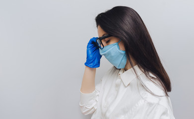 Young nurse doctor in a protective blue medical mask and gloves holding glasses in his hand and looking forward. Physical fatigue, sleepiness. Concept of quarantine, pandemic, coronavirus COVID-19.