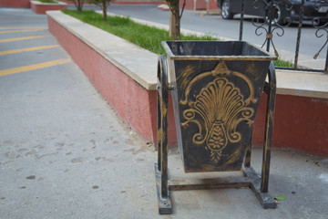 Black copper metal garbage bin in building with blurred . Garbage can on top of asphalt . Black trash can in the yard of the building .