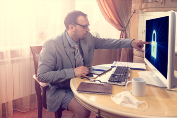 Sitting at the kitchen table, a young man in a business suit and shorts holds works remotely on...