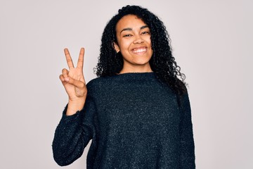 Young african american curly woman wearing casual sweater standing over white background smiling with happy face winking at the camera doing victory sign. Number two.