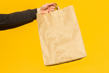 Close up male hold in hand brown empty blank craft paper bag for takeaway on yellow background. Packaging template mockup. food delivery service during coronavirus, concept. Copy space for Advertising