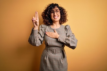 Beautiful arab business woman wearing dress and glasses standing over yellow background smiling swearing with hand on chest and fingers up, making a loyalty promise oath
