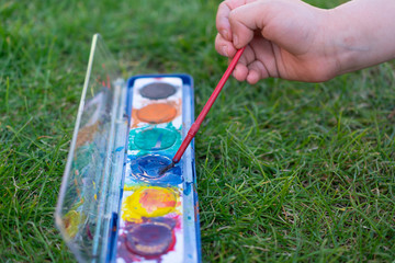 Child hand holding brush in watercolors paint set outdoors on green grass