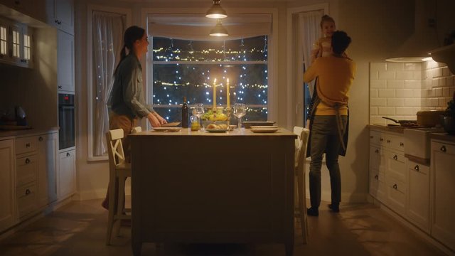 Family of Three Cooking and Having Dinner Together. Mother Prepares Food, Little Girl Runs to Father, Hugs Him and They Dance. Festive Table in Stylish Kitchen Interior with Warm Light. Slow motion 
