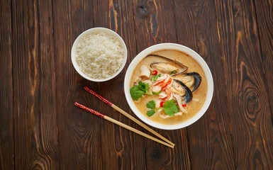 Tom yam spicy thai soup with shrimp, seafood, coconut milk and chili pepper in white bowl with rice and chopsticks over wooden texture background top view flat lay, copy space asian food concept