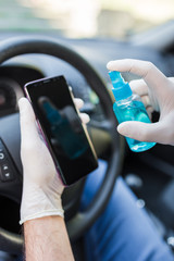 Close up of man driver wearing protective gloves and cleaning smartphone with alcohol spray in car. Germs or bacteria contamination.