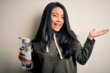 Young beautiful chinese sportswoman holding bottle of water over isolated white background very happy and excited, winner expression celebrating victory screaming with big smile and raised hands