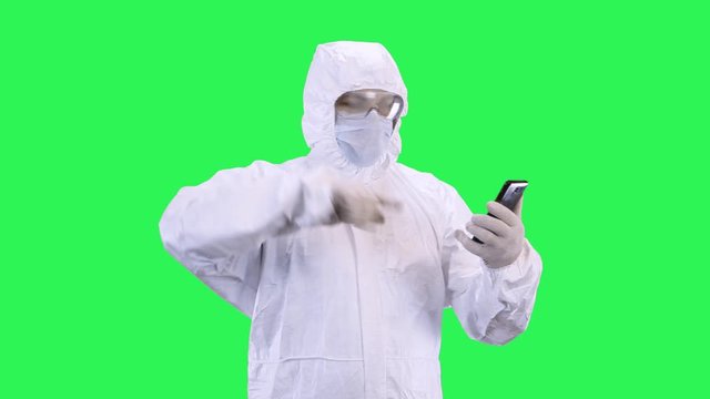 A man in a mask and a protective suit is talking via video calling on a smartphone while standing against a green background, quarreling and cursing with an interlocutor