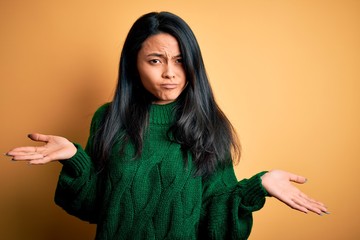 Young beautiful chinese woman wearing green sweater over isolated yellow background clueless and confused expression with arms and hands raised. Doubt concept.