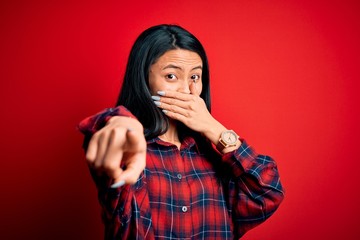 Young beautiful chinese woman wearing casual shirt over isolated red background laughing at you, pointing finger to the camera with hand over mouth, shame expression