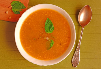 Top view of a plate of pumpkin soup with basil leaves and pine nuts on wooden plate and spoon on green background