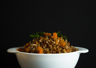 Buckwheat porridge with carrot slices and a parsley leaf in a white bowl close up on a black background with space