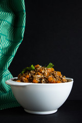 Buckwheat porridge with carrot slices and a parsley leaf in a white bowl and green textiles close up on a black background with copy space