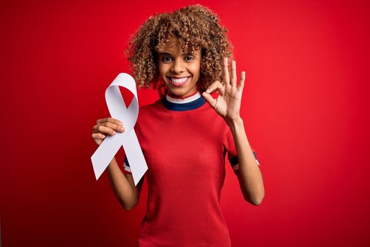 Young african american woman with curly hair holding white cancer ribbon over red background doing ok sign with fingers, excellent symbol