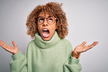 Young beautiful african american woman wearing turtleneck sweater and glasses crazy and mad shouting and yelling with aggressive expression and arms raised. Frustration concept.