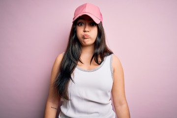 Young brunette woman wearing casual sport cap over pink background puffing cheeks with funny face. Mouth inflated with air, crazy expression.