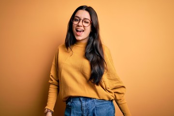 Young brunette woman wearing glasses and casual sweater over yellow isolated background winking looking at the camera with sexy expression, cheerful and happy face.
