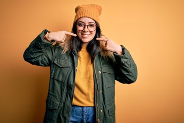 Young brunette woman wearing glasses and winter coat with hat over yellow isolated background smiling cheerful showing and pointing with fingers teeth and mouth. Dental health concept.