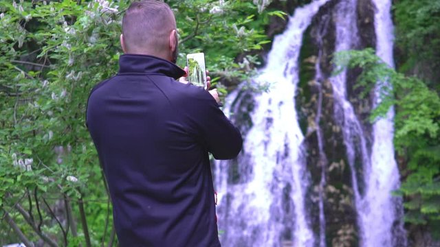 Man admiring the waterfall and doing photos on smartphone
