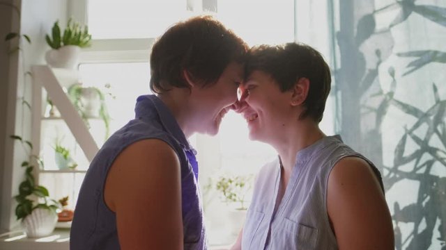 Two young women hug and kiss at home by the window. Casual clothing. Lesbian couple, homosexual relations, same-sex love.