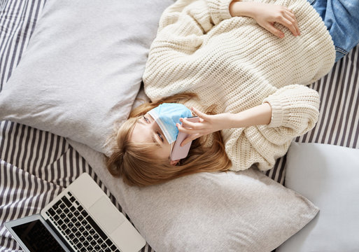 Young attractive woman in medical mask wearing white sweater and jeans on quarantine. Teen talking via cell phone lying on a bed with laptop nearby, wearing protective mask.