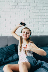 beautiful young Caucasian teenage girl sitting in bed with modern headphones is holding phone in her hand, dancing to cool music against large white wall. Active teenager morning. place for text
