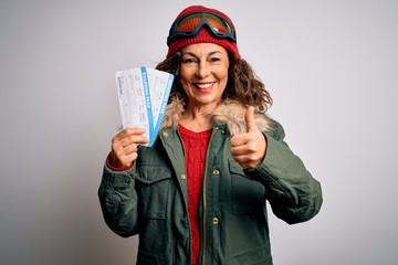 Middle age tourist woman wearing skier goggles holding airline boarding pass happy with big smile doing ok sign, thumb up with fingers, excellent sign