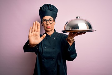 Middle age brunette chef woman wearing cooker uniform and hat holding tray dome with open hand doing stop sign with serious and confident expression, defense gesture