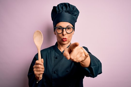 Middle age brunette chef woman wearing cooker uniform and hat holding wooden spoon pointing with finger to the camera and to you, hand sign, positive and confident gesture from the front