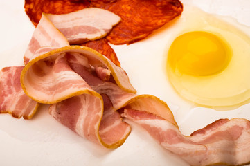 A broken chicken egg without a shell, slices of sausage and bacon and tomatoes on a white background. Close up.