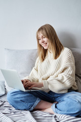Happy attractive young girl sitting on the sofa and typing on laptop, enjoying writing blog or chatting with friends in social network. Concept of communicating, working or studying online