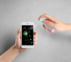 Woman sanitizing smartphone with antiseptic spray over grey table, closeup. Be safety during coronavirus outbreak