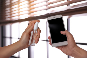Woman sanitizing smartphone with antiseptic spray indoors, closeup. Be safety during coronavirus outbreak