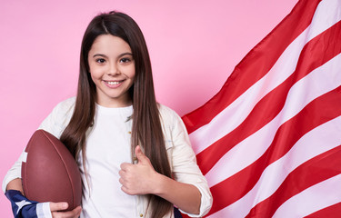 The American sport football.USA. Young beautiful girl in a white T-shirt with the American flag, on a pink background.