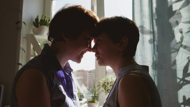 Two young women hug and kiss at home by the window. Casual clothing. Lesbian couple, homosexual relations, same-sex love.