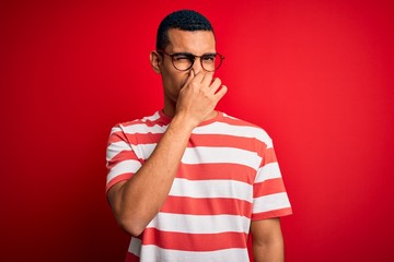Young handsome african american man wearing casual striped t-shirt and glasses smelling something...