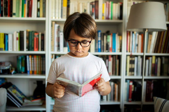 Portrait of serious boy with glasses standing in front of book shelves reading a comic