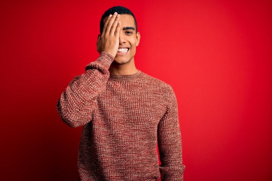Young handsome african american man wearing casual sweater standing over red background covering one eye with hand, confident smile on face and surprise emotion.
