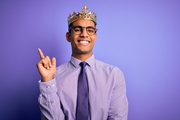 Young handsome african american man wearing golden crown of king over purple background with a big smile on face, pointing with hand and finger to the side looking at the camera.