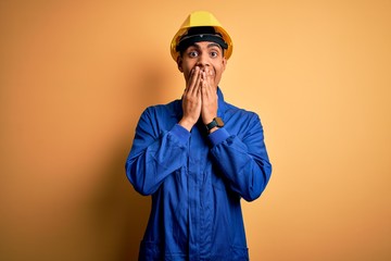 Young handsome african american worker man wearing blue uniform and security helmet laughing and embarrassed giggle covering mouth with hands, gossip and scandal concept